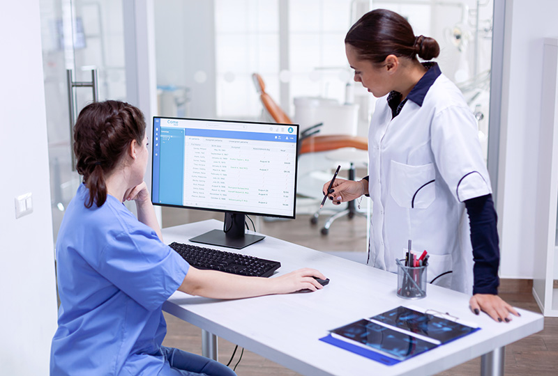 A woman in a white lab coat stands at a desk with a computer monitor, interacting with a patient seated across from her.