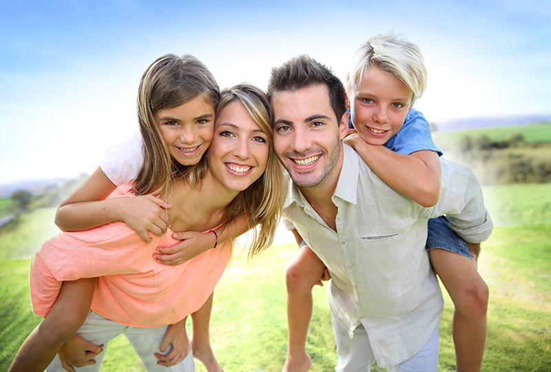 Family of four with a smiling man carrying two children, posing outdoors on grass.