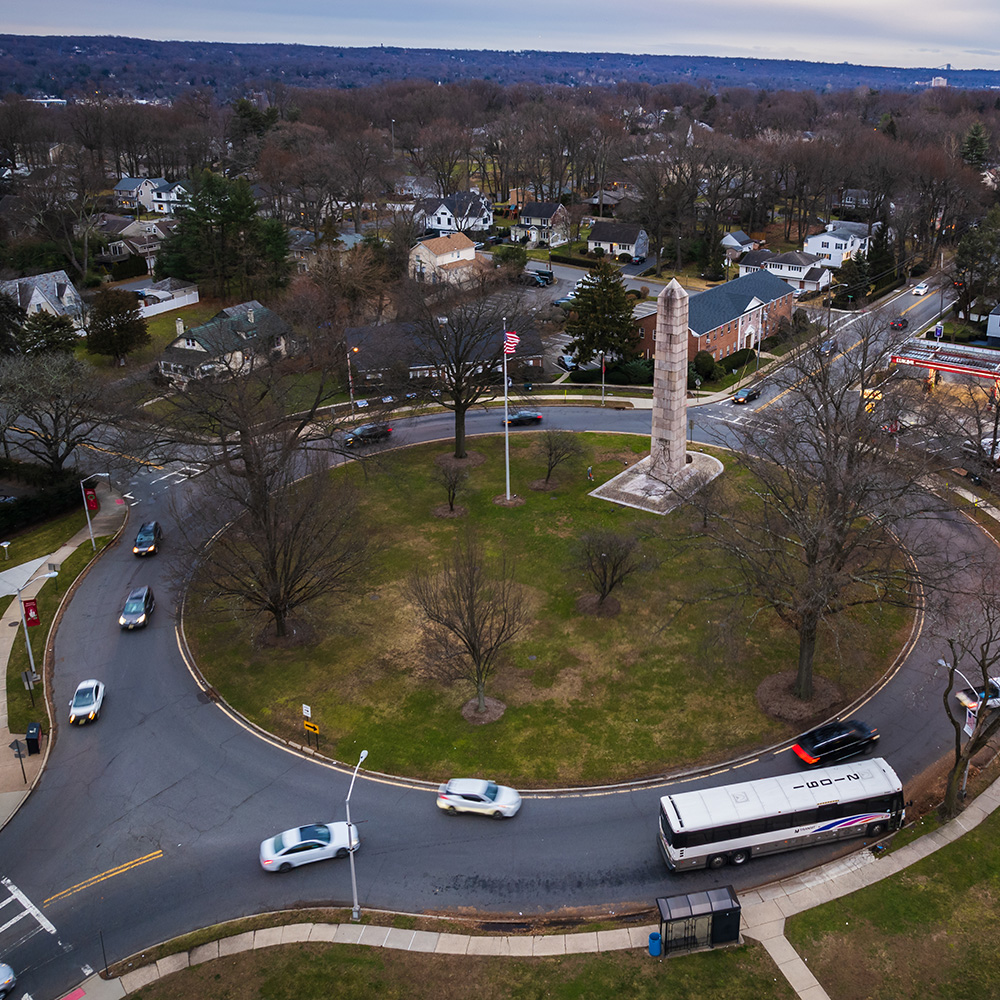 A roundabout in a suburban area with vehicles and traffic lights, surrounded by grass and trees.